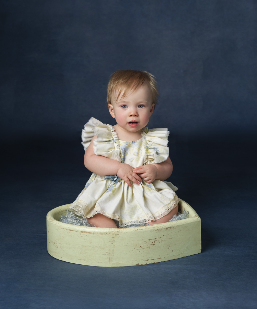 one year old sitting session of a little girl wearing a dress sitting in a heart bowl with bright blue eyes